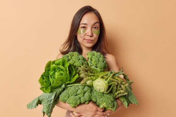 asian woman with dark hair looks seriously at camera raises eyebrows carries big heap of fresh vegetables applies hydrogel patches under eyes to reduce wrinkles poses against beige background