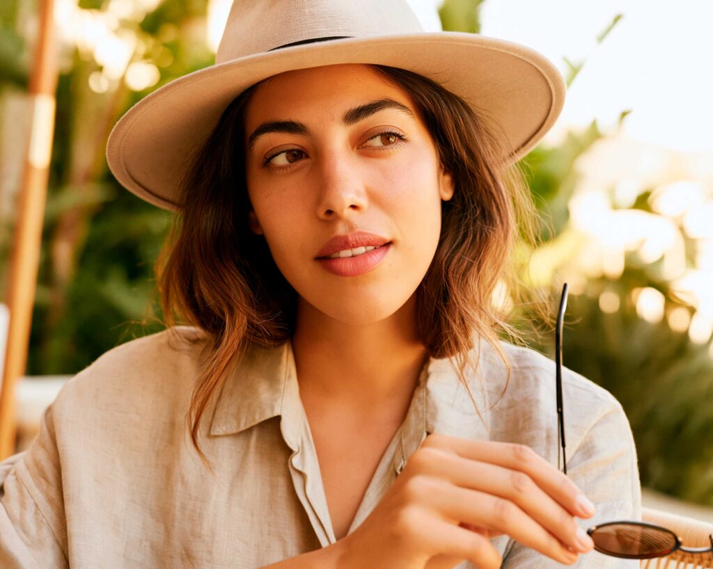 Woman in beige hat holding sunglasses outdoors