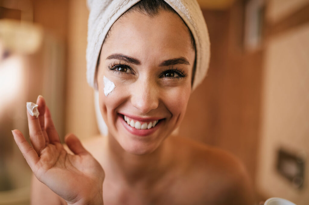 Smiling woman applies face cream with towel-wrapped hair