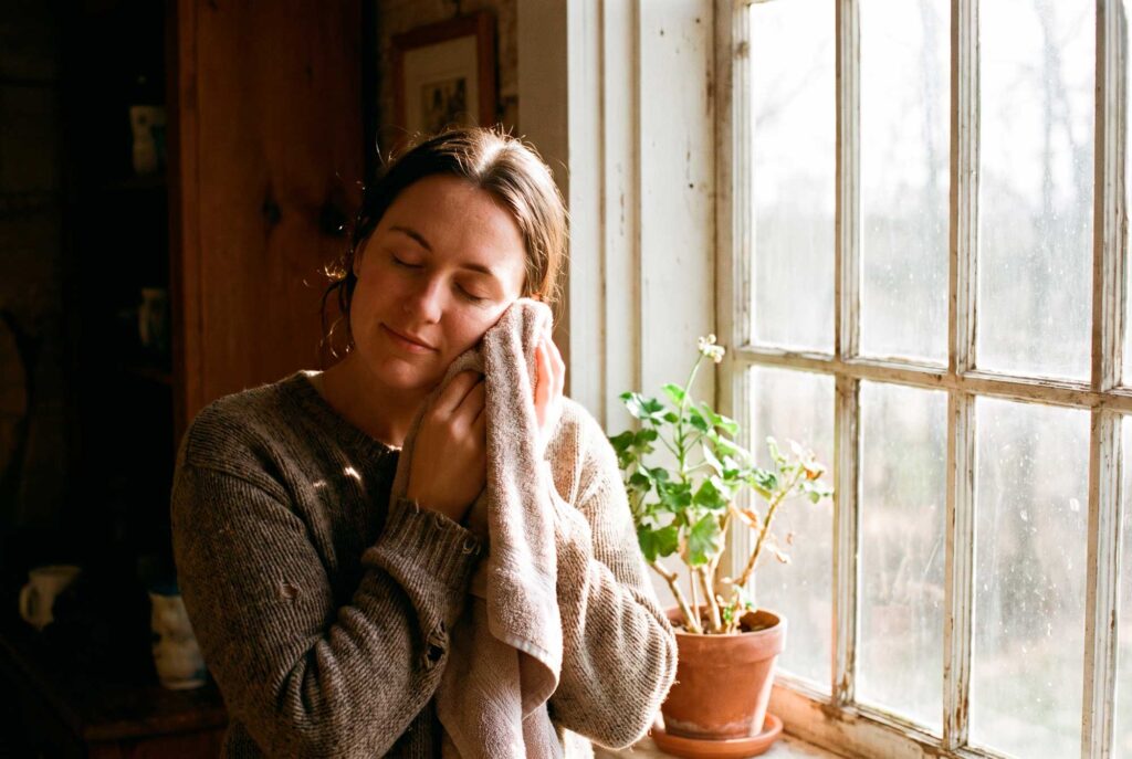 Woman holds towel to face beside sunny window